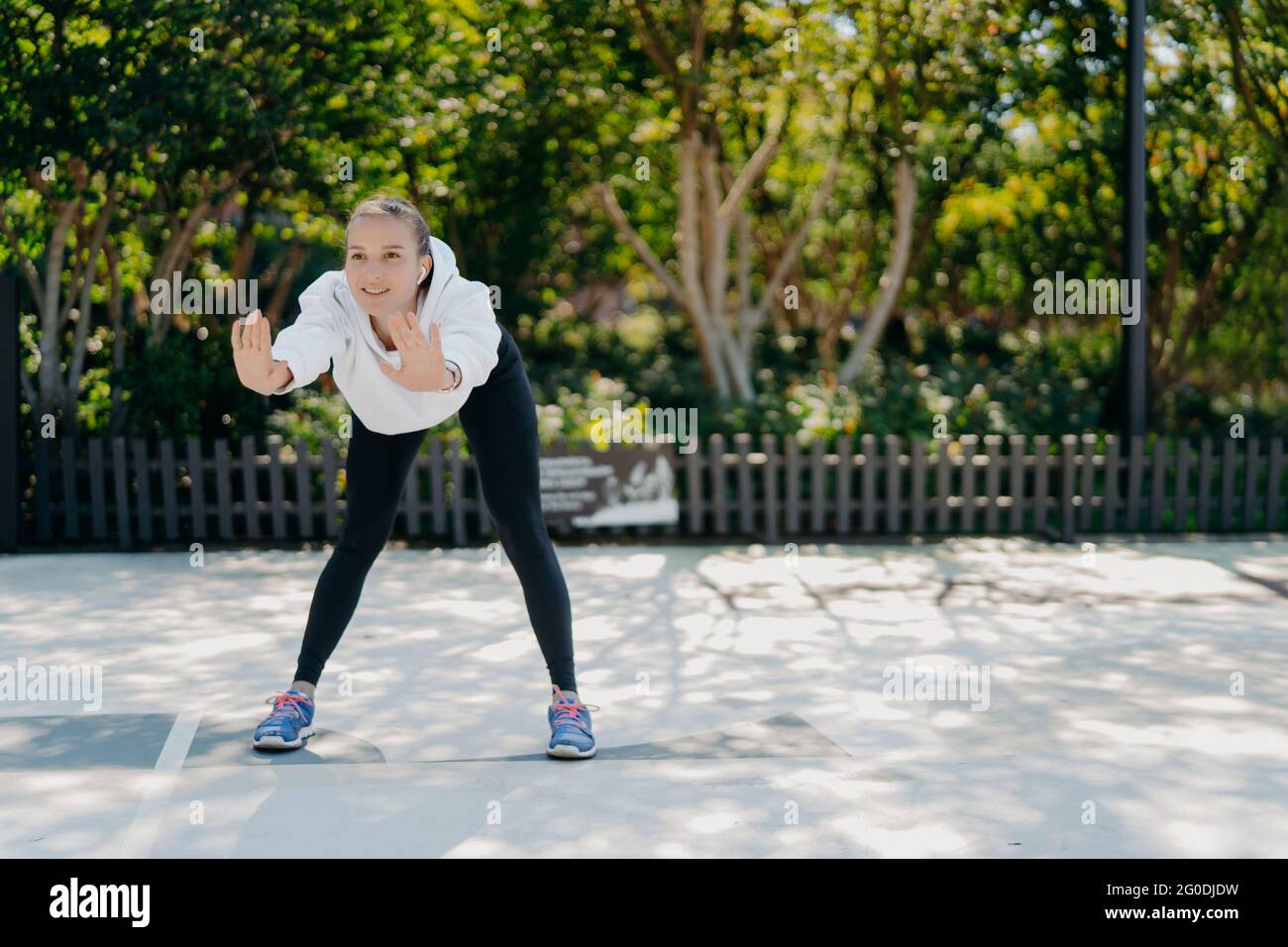 Zufriedene gesunde sportliche Frau lehnt sich nach vorne hält die Handflächen angehoben macht Übungen in Freiluftzügen im Park fühlt sich gut trägt Sweatshirt Leggings und Stockfoto