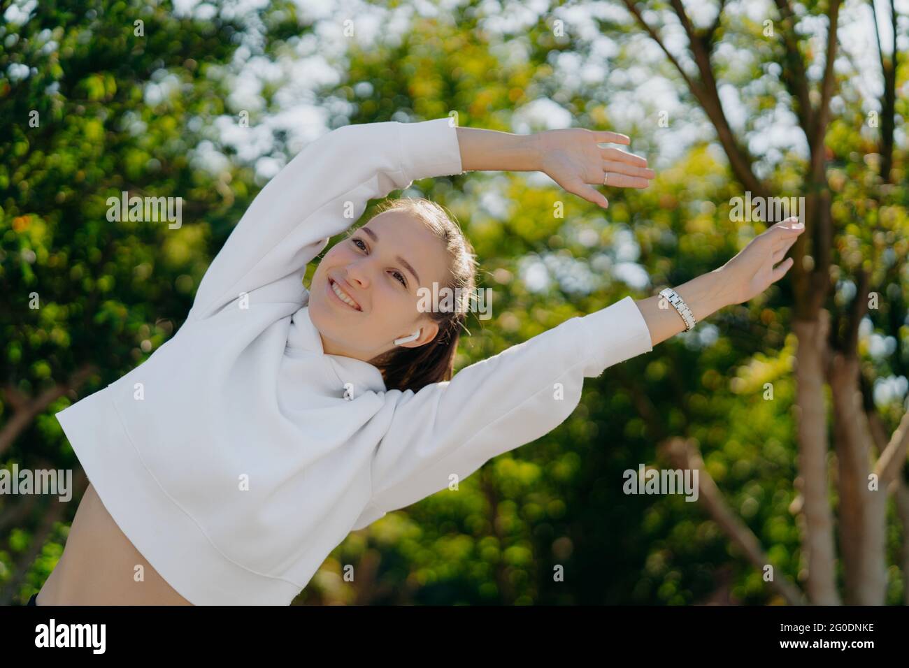 Aktive junge Frau streckt die Arme lehnt sich zur Seite lächelt positiv genießt das Training an der frischen Luft trägt weißes Sweatshirt hat regelmäßige Trainings oder Workout Stockfoto