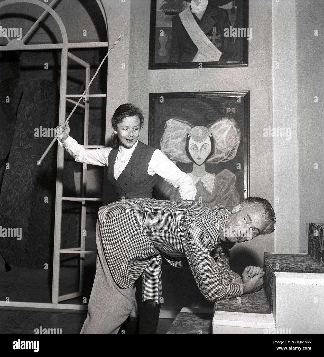 Hinter den Kulissen des Theaters in den 1950er Jahren. Die junge Schauspielerin Elisaveta, 1922-1992, hält einen Stock in der Hand, während der Schauspieler Egon Larsson sich nach vorne gebeugt hat, um seine Strafe zu erhalten. Einfach nur lustig und nicht wirklich. Schweden 1953 Ref. BM28-11 Stockfoto