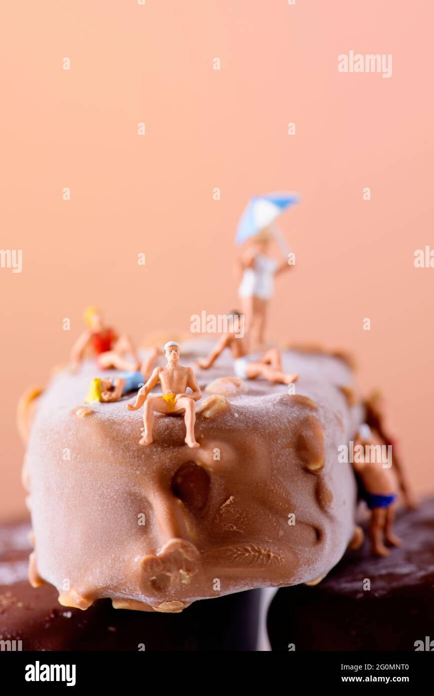 Eine Gruppe von Miniatur-Männern und -Frauen, die Badeanzug tragen, und eine alte Dame, die einen Regenschirm trägt, sich an einigen Schokoladen-Eisdielen entspannt, gegen ein Braun Stockfoto