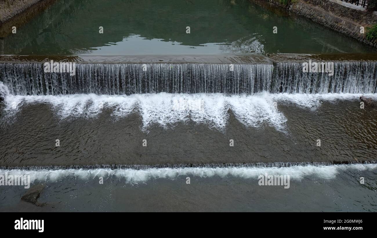 Ein breiter, aber flacher Fluss mit einer Betonwand auf beiden Seiten und einer Betonwand, die in der Mitte einen kleinen Wasserfall bildet. Stockfoto