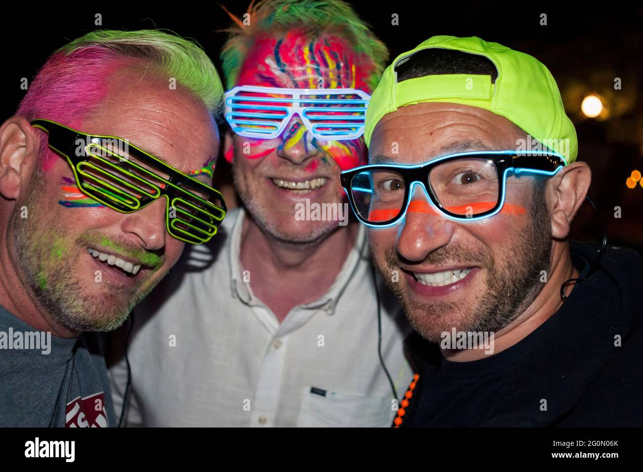 Eine Gruppe von drei Männern an einem Abend, die auf einem Festival mit Neon-Gesichtsfarbe feiern und LED-Lichtbrillen aufleuchten Stockfoto