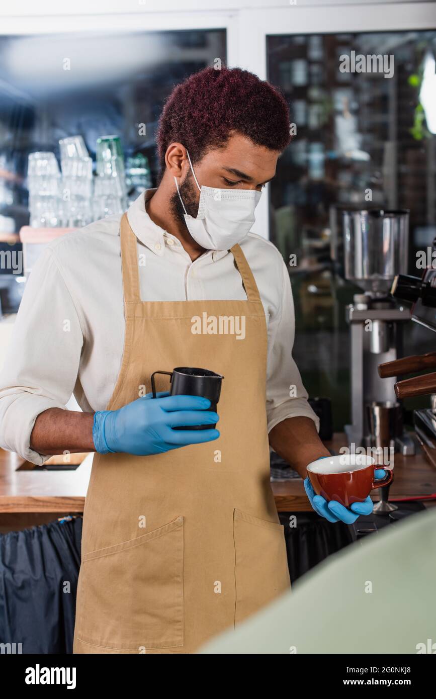 afroamerikanischer Barista in medizinischer Maske mit Tasse und Milchkrug in der Nähe der Kaffeemaschine Stockfoto