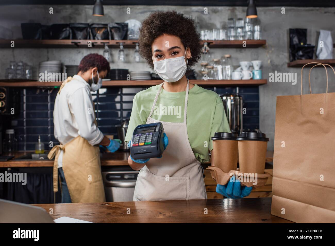 afroamerikanischer Barista in medizinischer Maske und Latexhandschuhen mit Pappbechern und Zahlungsterminal Stockfoto