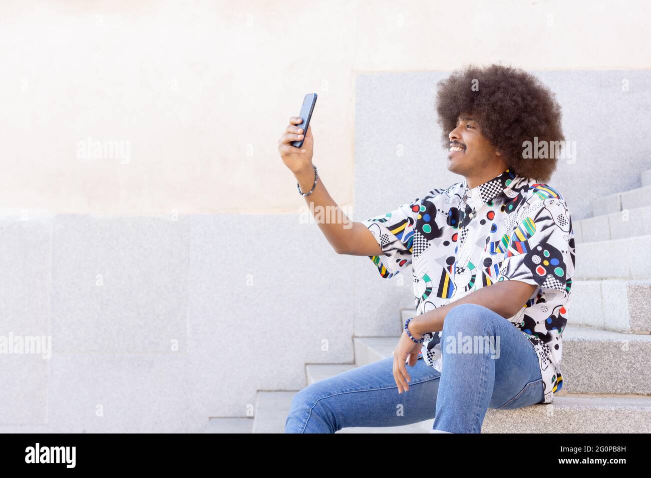Schwarzer Kerl mit afro Haaren lächelnd sitzt auf einigen Steinstufen und nimmt ein Selfie mit seinem Smartphone Stockfoto