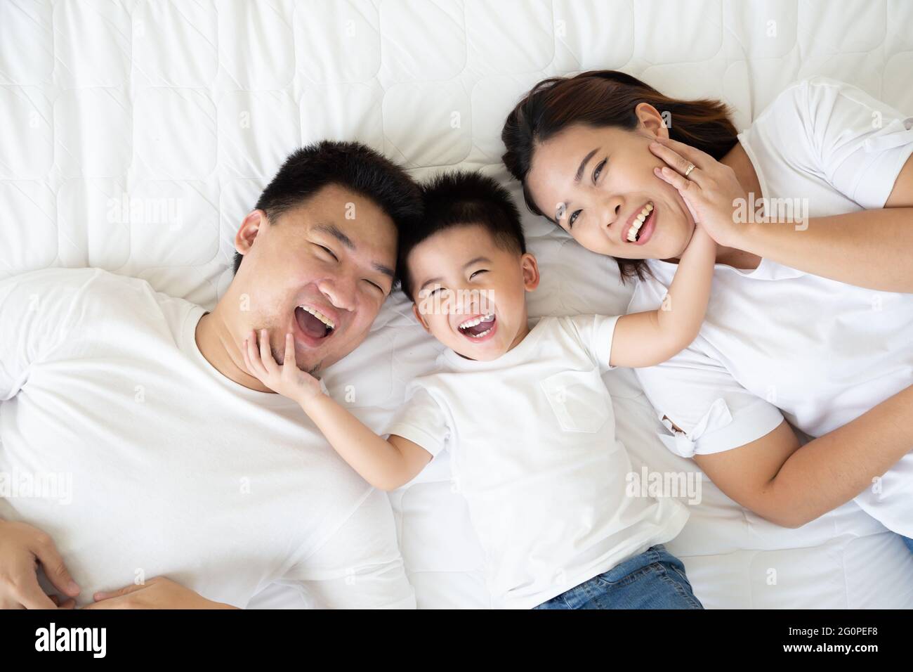 Draufsicht auf den niedlichen kleinen Jungen und seine asiatischen Eltern, die die Kamera betrachten und lächeln, während sie zu Hause auf dem weißen Bett liegen, Happy Family Konzept Stockfoto