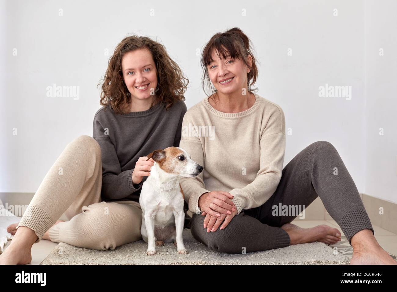 Fröhliche Erwachsene Tochter und Mutter mittleren Alters sitzen auf dem Boden Mit niedlichem Hund zu Hause Stockfoto