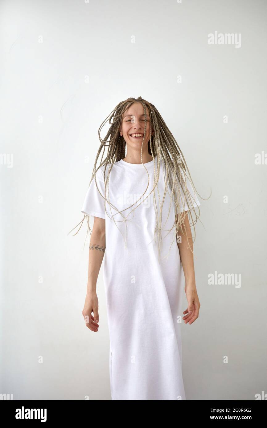 Fröhliche junge Hündin mit Zöpfen im lässigen maxi-weißen Kleid, das im Studio gegen eine helle Wand steht Stockfoto