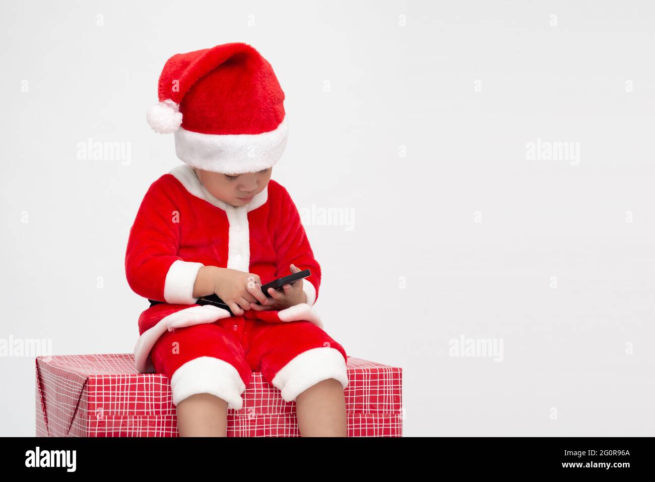 Asiatische Baby mit Weihnachtsmann Anzug halten Handy und sitzt auf Geschenk-Box isoliert auf weißem Hintergrund Stockfoto