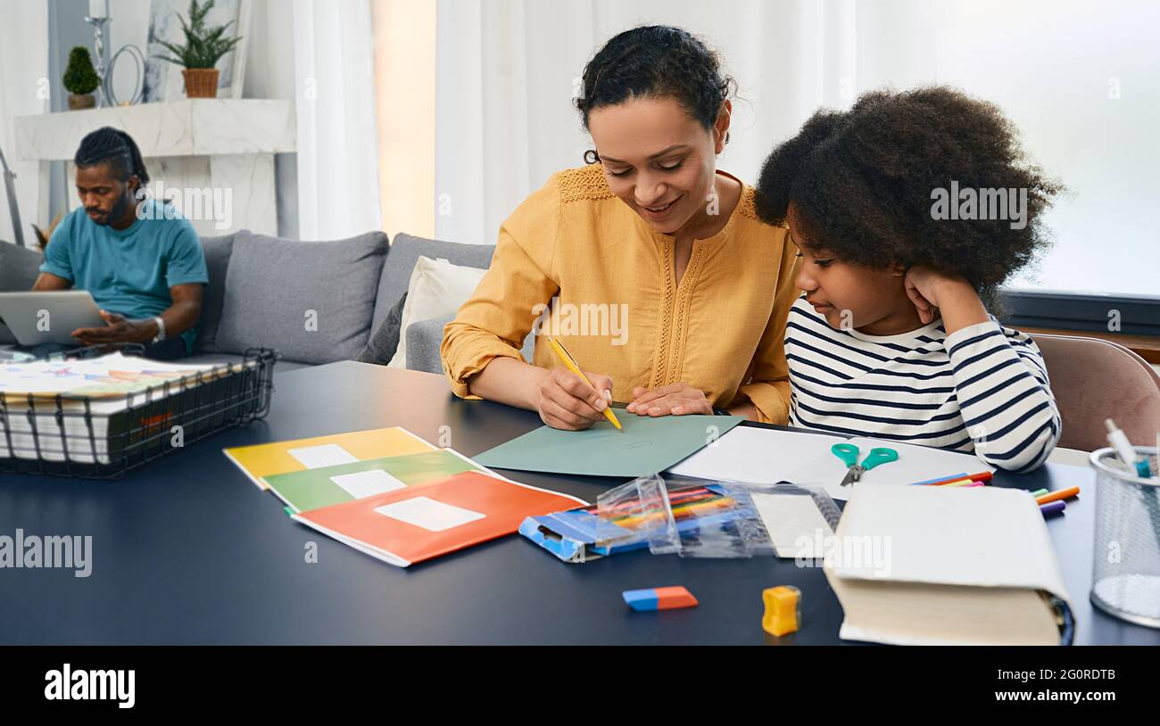 Familientag. Mutter hilft ihrer Tochter bei den Schulaufgaben, während der Vater an seinem Laptop arbeitet. Glückliche Familie, Komfort zu Hause Stockfoto