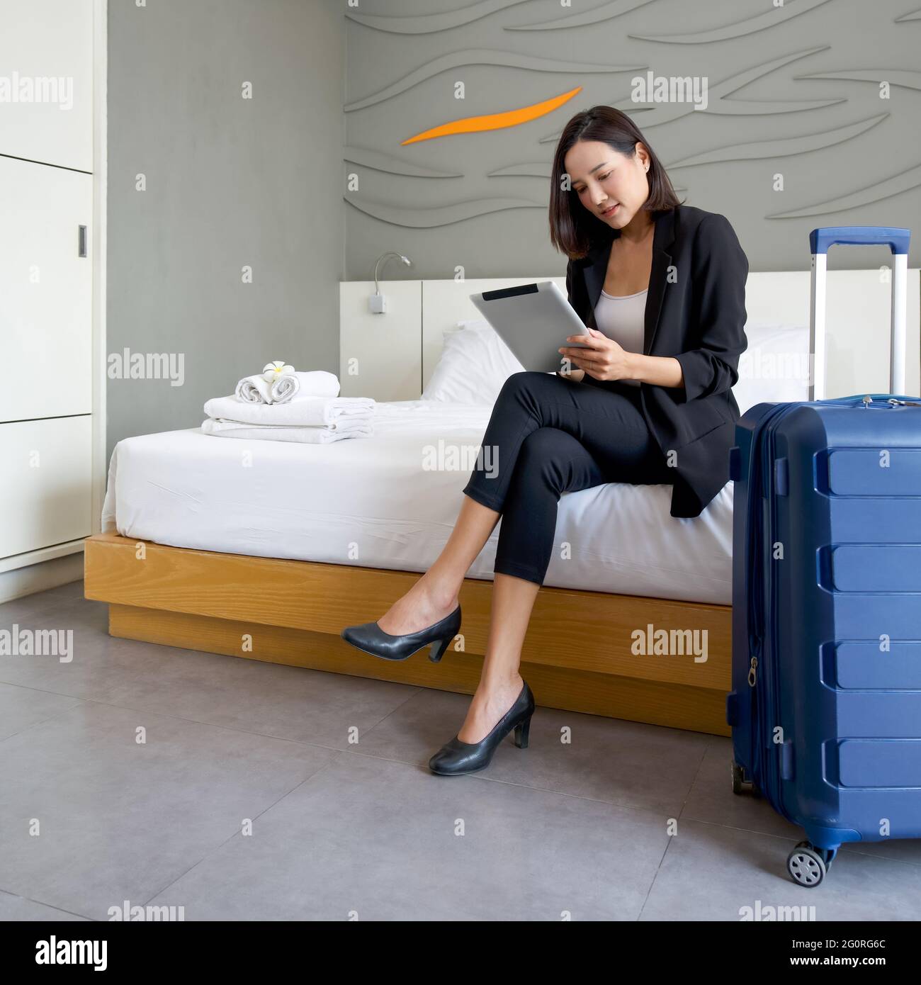 Asiatische Geschäftsfrau in schwarzem Anzug sitzt mit Tablet-Computer in  der Hand. Ihr Gepäck wurde neben das Bett gelegt. Die Atmosphäre der Arbeit  im Freien während des Th Stockfotografie - Alamy