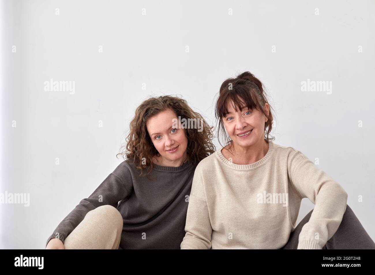 Fröhliche Erwachsene Tochter und Mutter mittleren Alters sitzen auf dem Boden zu Hause auf weißem Hintergrund Stockfoto