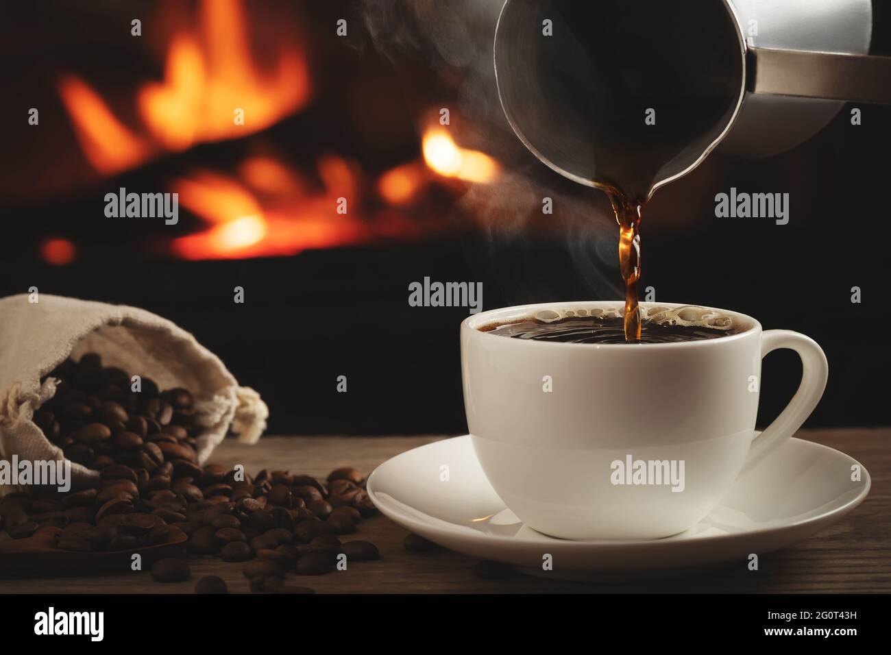 Gießen von Cezve-Kaffee in eine Tasse auf einem Holztisch vor einem brennenden Kamin. Selektiver Fokus. Stockfoto