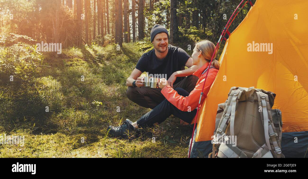 Camping im Freien - junges Paar, das sich nach dem Wandern bei einem Zelt mit Teetassen im Waldcamp ausruhen kann. Aktiver Lebensstil Stockfoto
