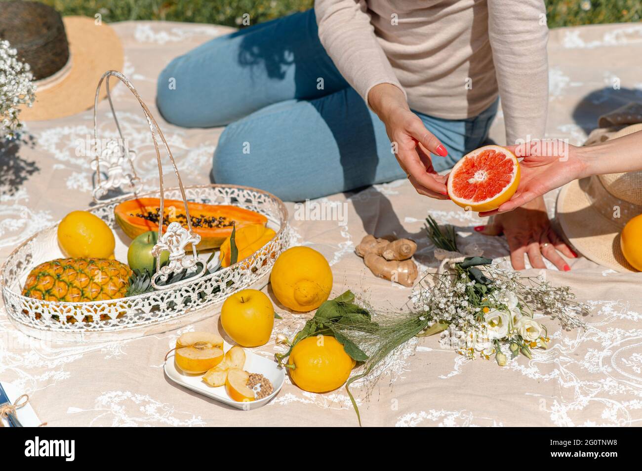 Unkenntliche Frauen sitzen auf einer Decke und picknicken. Weibchen, die eine rosa Grapefruit passieren. Vielfalt an tropischen Früchten in der Umgebung. Stockfoto