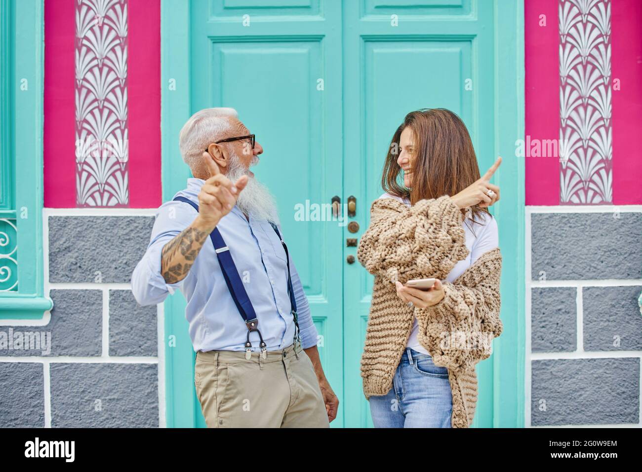 Eine glückliche Frau mittleren Alters teilt Daten in sozialen Medien mit einem stilvollen älteren Mann, während sie auf der Straße durch ein Mobiltelefon gegen ein farbenfrohes Gebäude surft Stockfoto