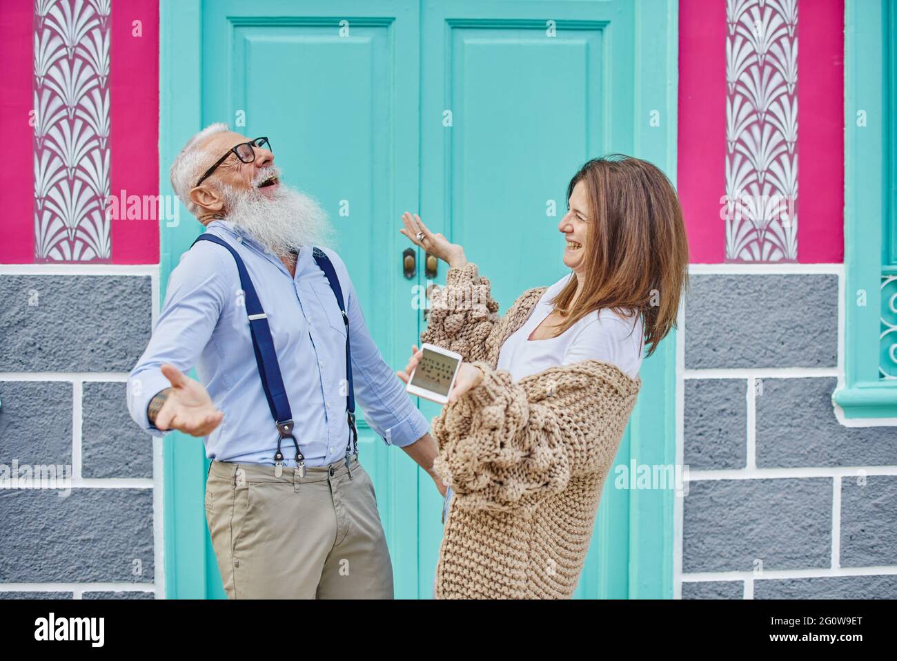 Eine glückliche Frau mittleren Alters teilt Daten in sozialen Medien mit einem stilvollen älteren Mann, während sie auf der Straße durch ein Mobiltelefon gegen ein farbenfrohes Gebäude surft Stockfoto