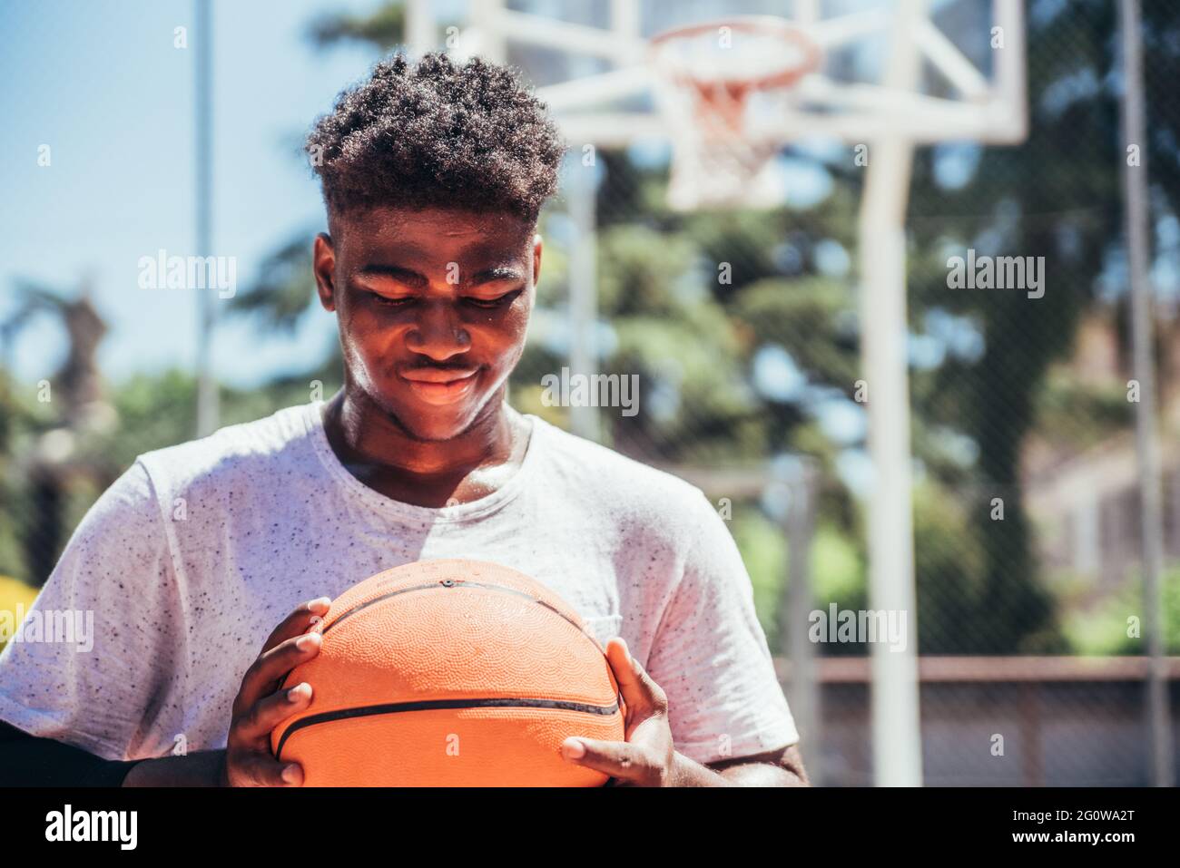 Porträt eines schwarzen afroamerikanischen Jungen, der auf einem städtischen Basketballfeld einen Basketball gegen seine Brust hält. Stockfoto