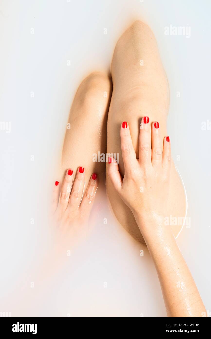 Detail der Hände einer Frau mit rot lackierten Nägeln, die auf ihren Beinen ruht, während sie in einer Badewanne voller Milch ein Bad nimmt. Körperpflegekonzept. Stockfoto