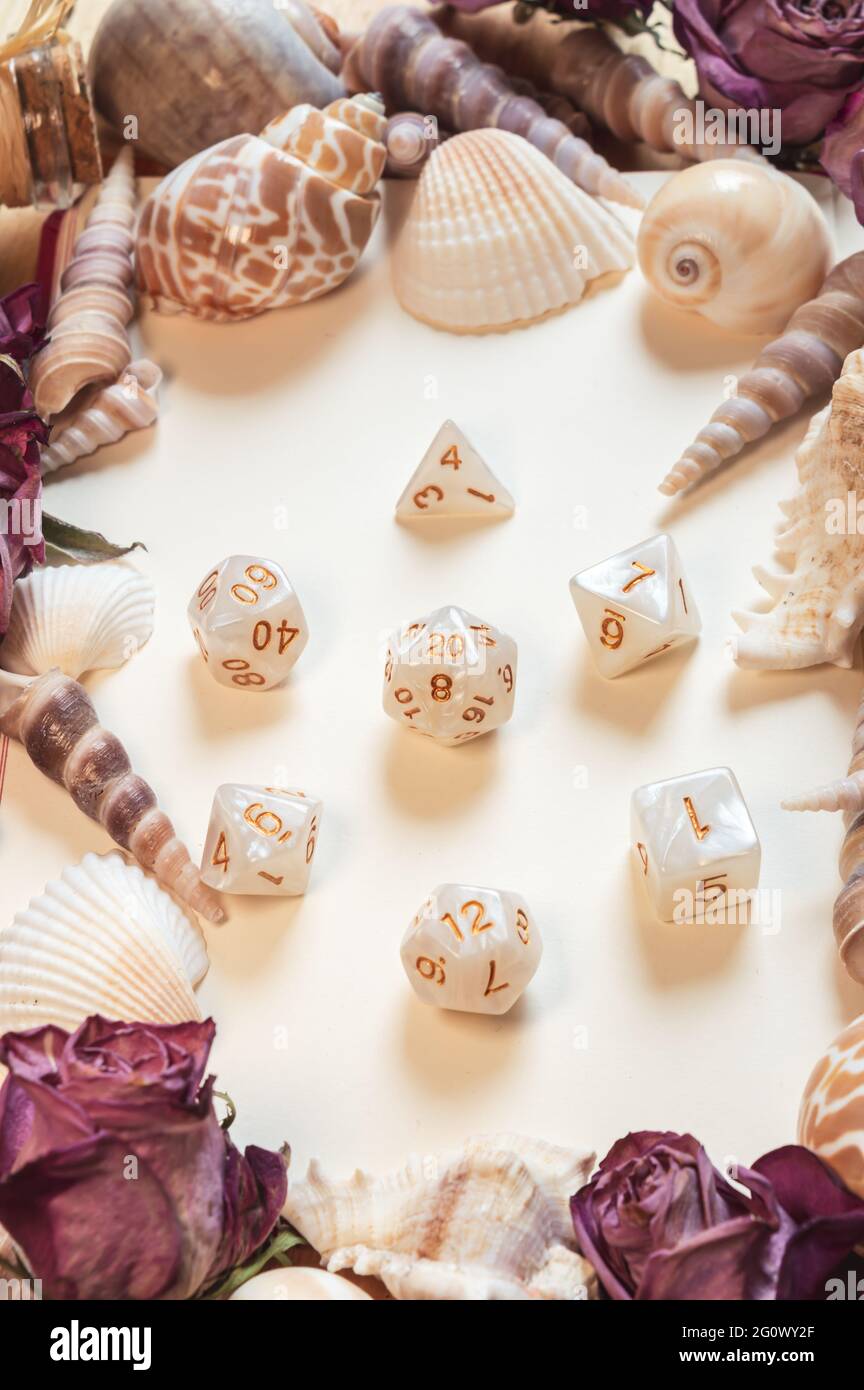 Vertikales Bild einer Reihe von weißen und goldenen RPG-Spielwürfeln, eingerahmt von Muscheln und getrockneten Rosen Stockfoto