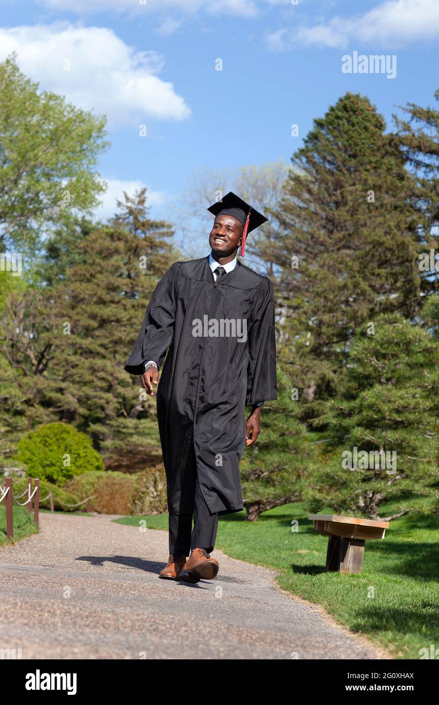 Ein junger, schwarzer Hochschulabsolvent mit Mütze und Kleid lächelt, als er stolz einen Pfad entlang geht Stockfoto