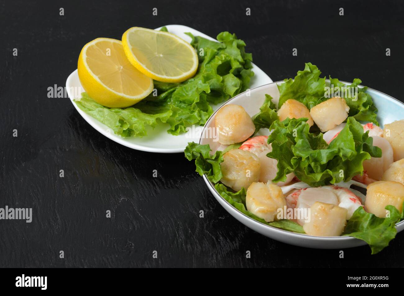 Gebratener Jakobsmuschel mit gekochtem Garnelenschwanz mit Salatblättern und Zitrone in einer Keramikplatte auf dunklem Hintergrund. Selektiver Fokus. Stockfoto
