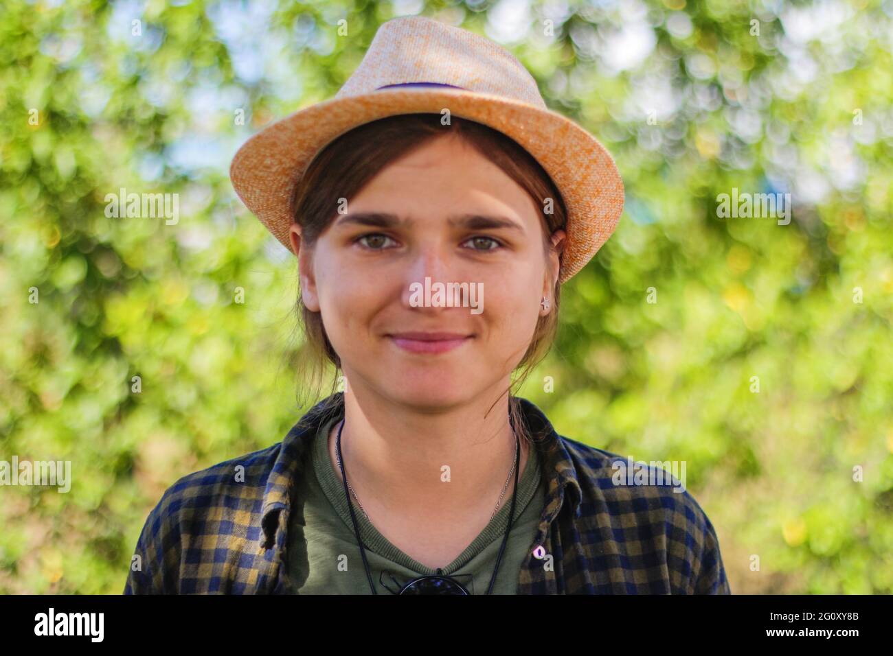 Defokussed Nahaufnahme Licht lächelnd junge Brünette kaukasische Frau in Hut. Sommer verschwommen grün Natur Bokeh Hintergrund. Porträt von schönen jungen niedlich Stockfoto