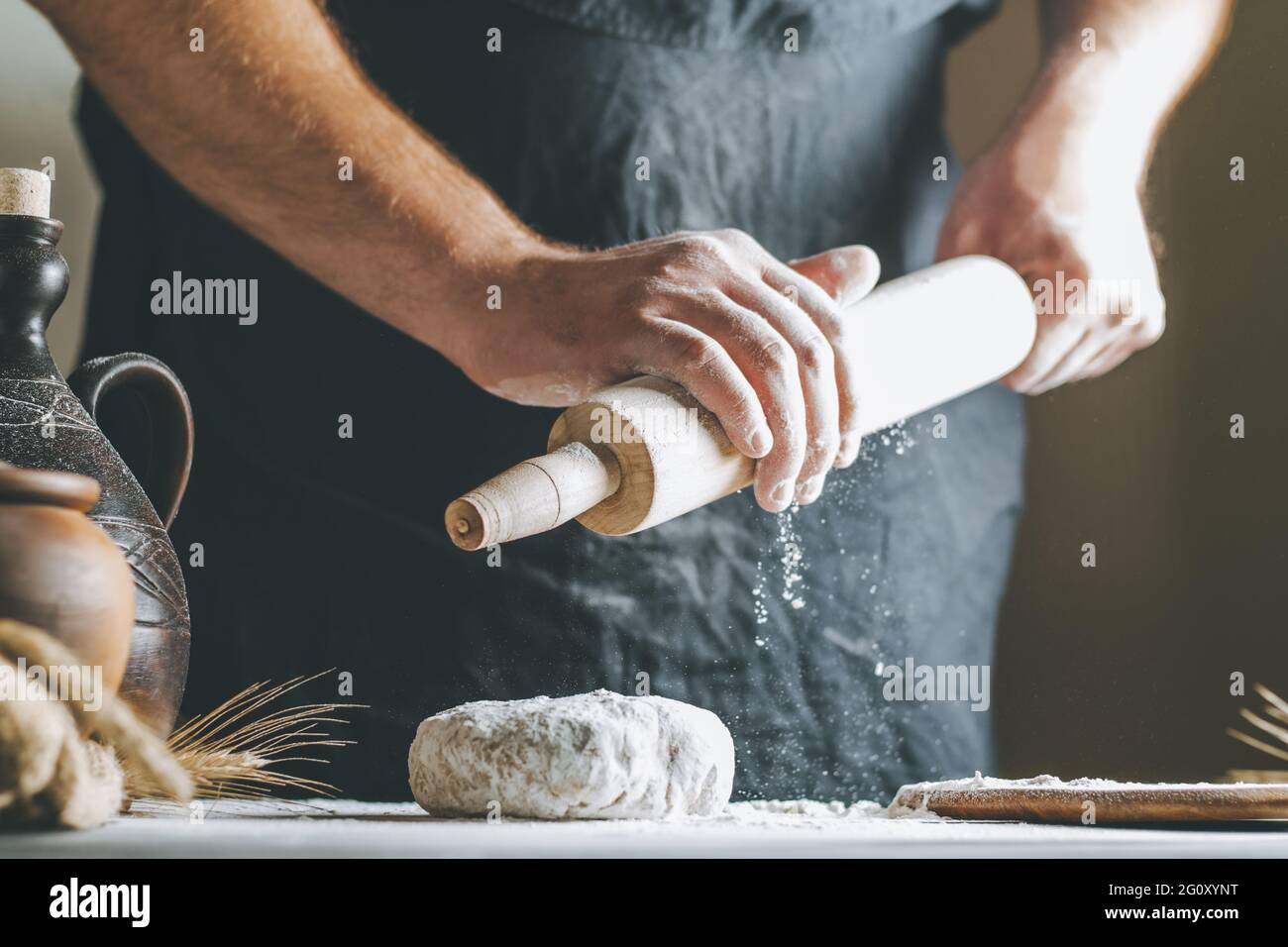 Männliche Hände gießen beim Kochen Mehl auf ein Nudelholz neben Teig, Tontopf, Ölflasche und Nudelholz auf einem dunklen Tisch Stockfoto