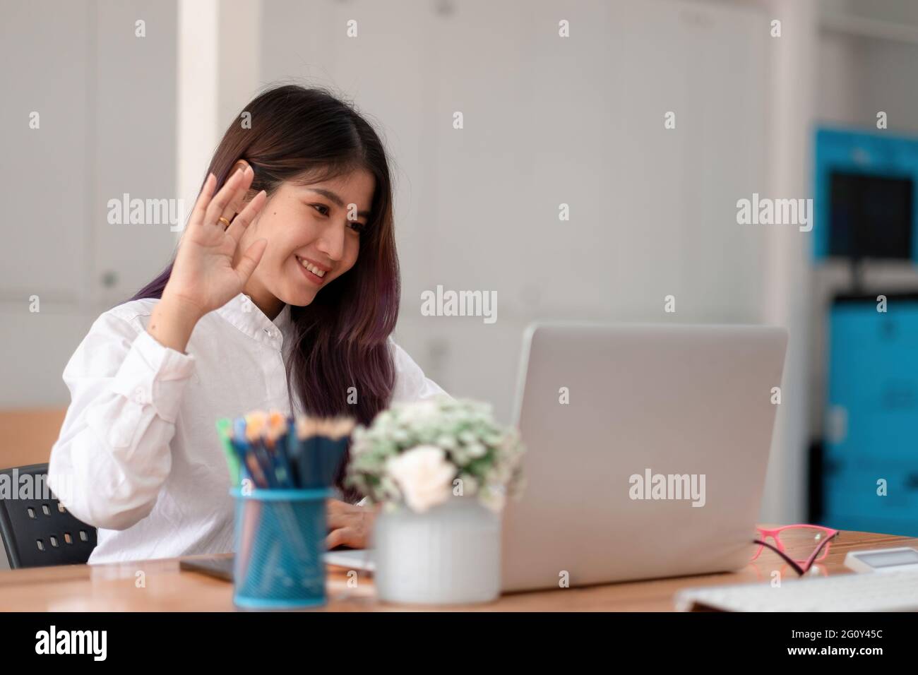 Bild einer glücklichen asiatischen Frau, die im weißen Hemd lächelt und die Hand am Laptop schwenkt, während sie im Büro spricht oder chattet Stockfoto