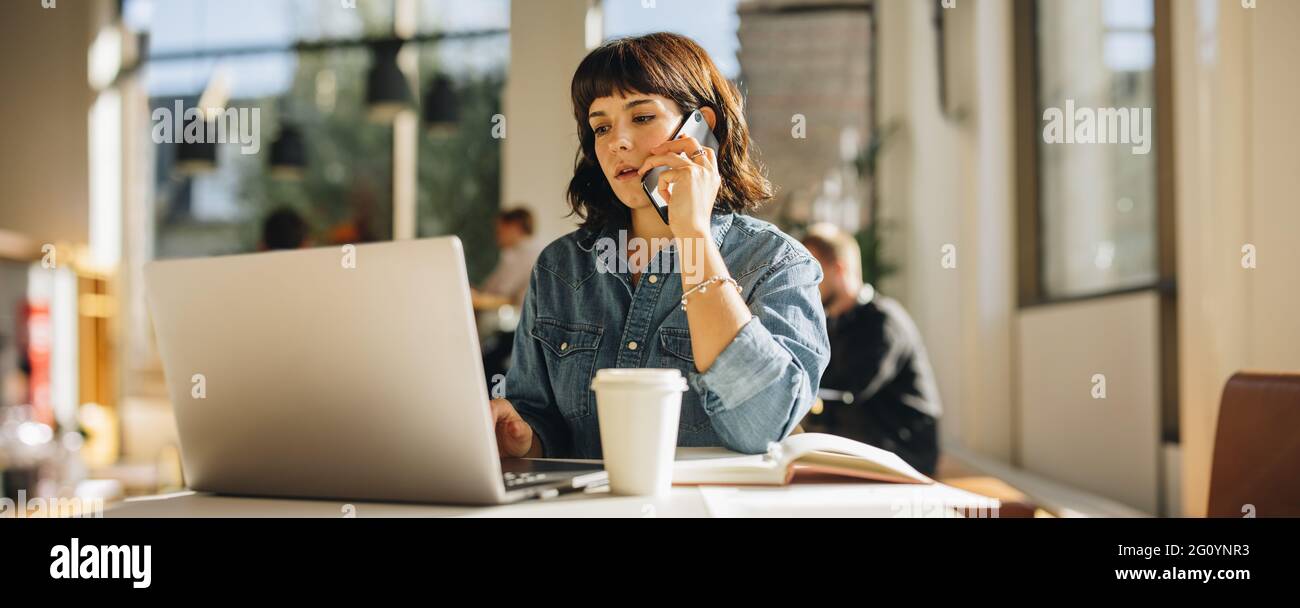 Junge Frau, die am Schreibtisch sitzt und auf dem Handy spricht und am Laptop arbeitet. Geschäftsfrau, die mit dem Smartphone spricht, während sie im Coworkin einen Laptop benutzt Stockfoto