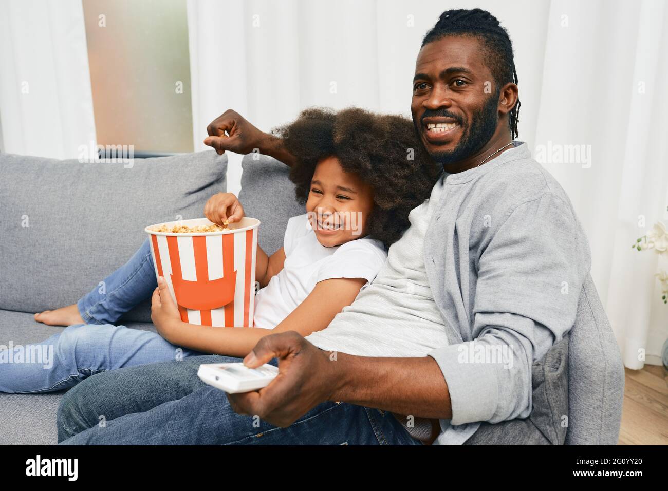Afroamerikanischer Vater mit seiner kleinen Tochter, die sich umarmt und gemeinsam Kino anschaut und Popcorn aus einem großen gestreiften Eimer isst. Vatertag Stockfoto