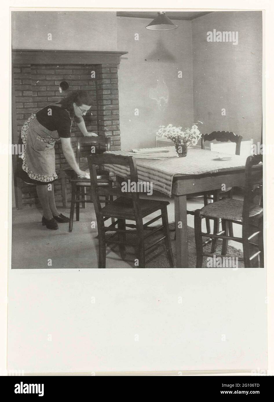 Frau aus Stoff. In einem schlicht eingerichteten Wohnzimmer staubt eine Frau einen Stuhl. Sie trägt eine Brille und eine karierte Schürze. Im Wohnzimmer gibt es einen Tisch mit einer karierten Tischdecke und einer Vase mit Blumen, eine Lampe an der Decke. Auf der Rückseite des Zimmers ein Kinderbett. Stockfoto