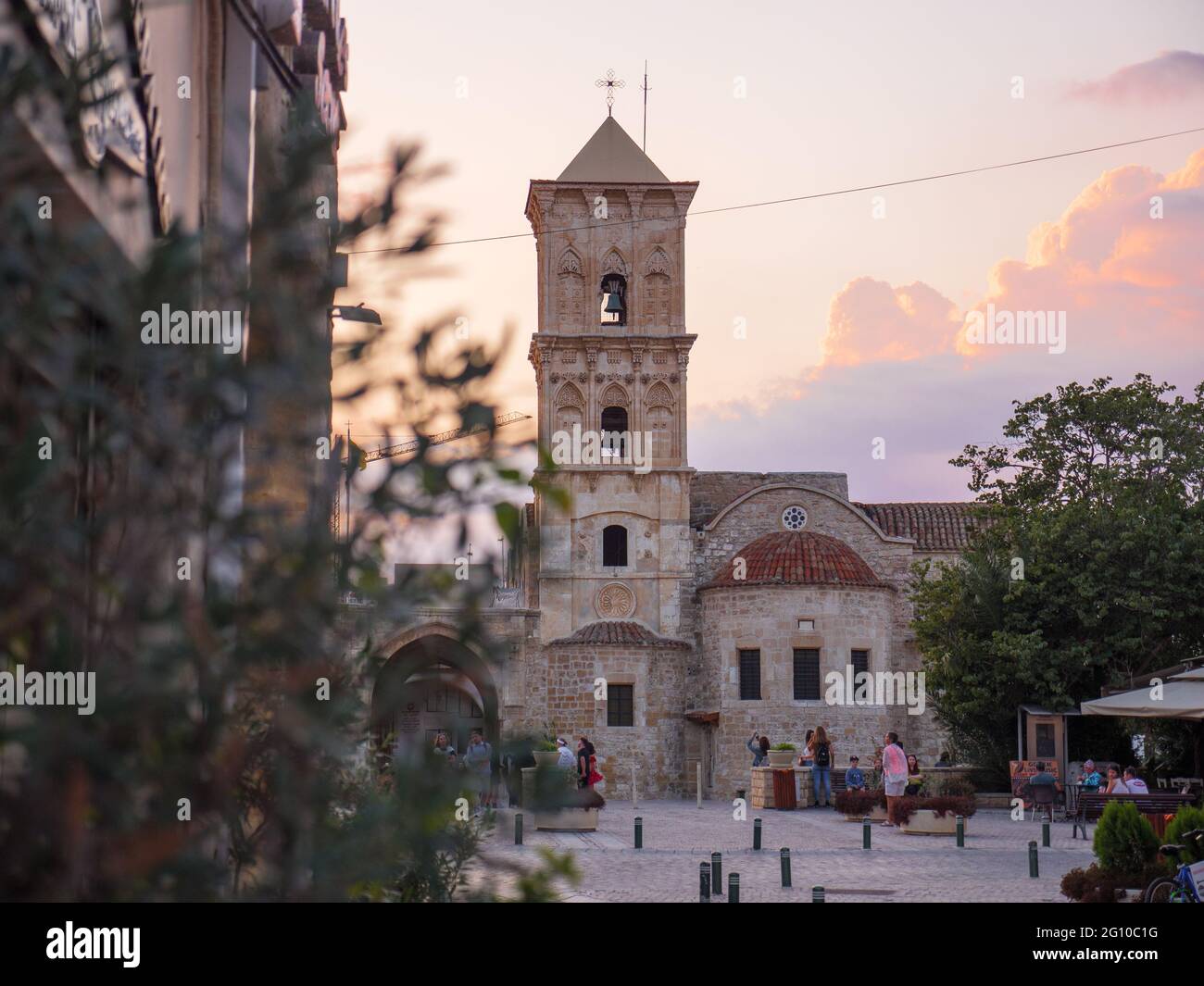 Blick durch Baumzweige über die st. Lazarus Kirche und Touristen sitzen und gehen auf dem Platz in der Nähe in Larnaca, Zypern. Historisches Erbe. Stockfoto