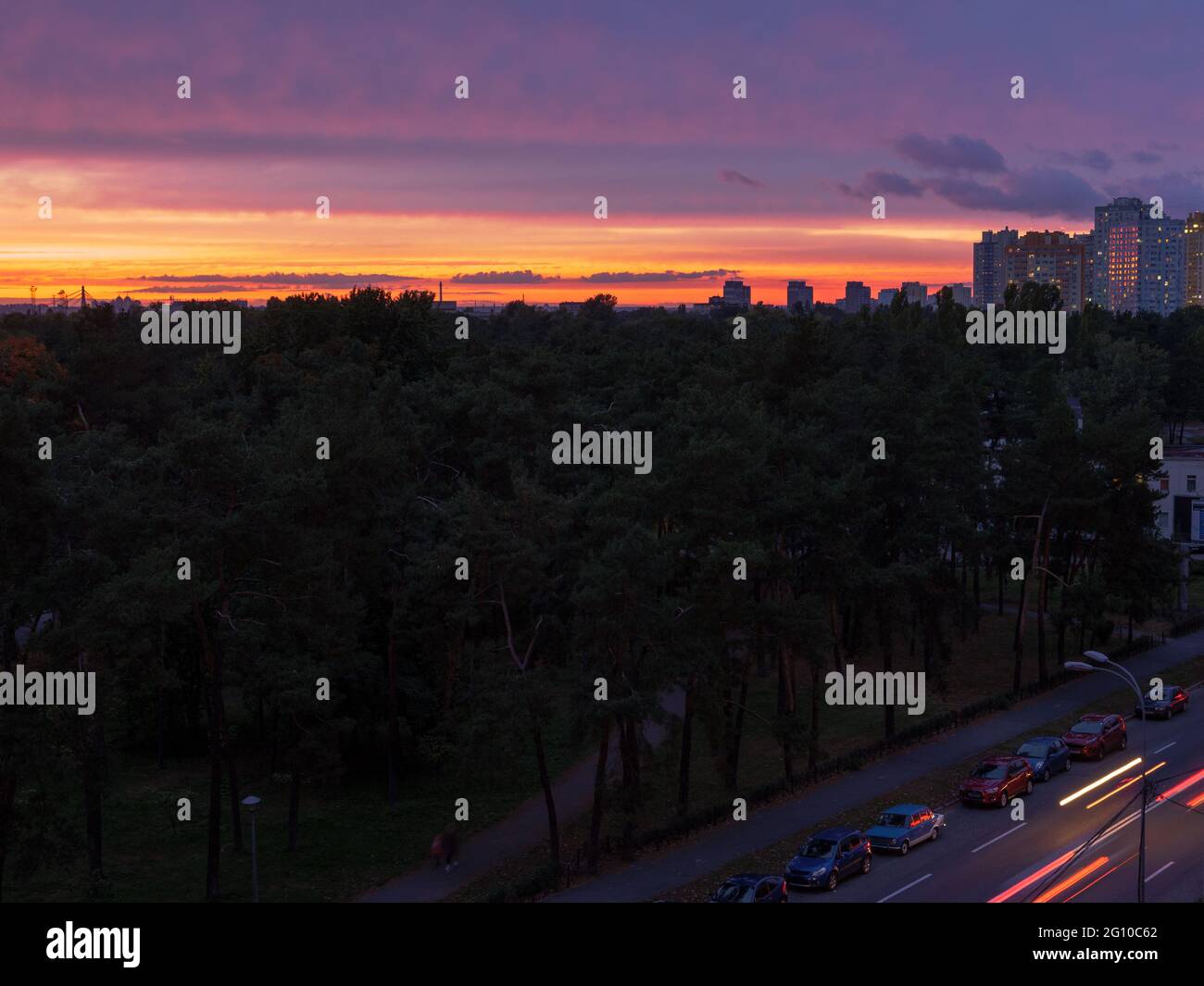 Erstaunlicher violetter, roter und gelber Sonnenuntergang über Kiew (Kiew) Stadt mit Gebäuden mit Licht in den Fenstern, Brücke und Straße mit Autos, die sich mit Scheinwerfern bewegen. Stockfoto
