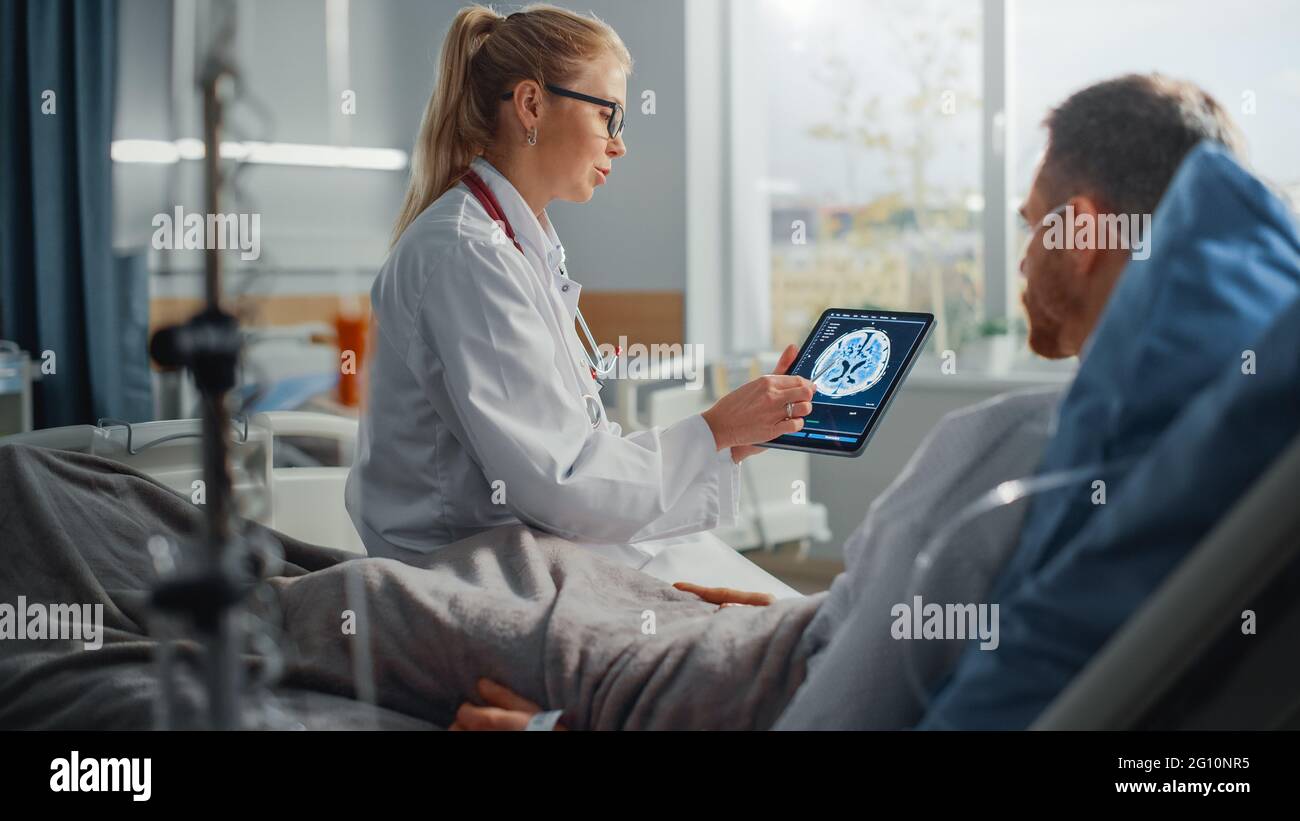 Krankenhausabteilung: Freundliche Ärztin spricht mit kranken männlichen Patienten, die sich im Bett ausruhen, verwendet einen Tablet-Computer, zeigt und erklärt die Ergebnisse von Gehirnscans Stockfoto