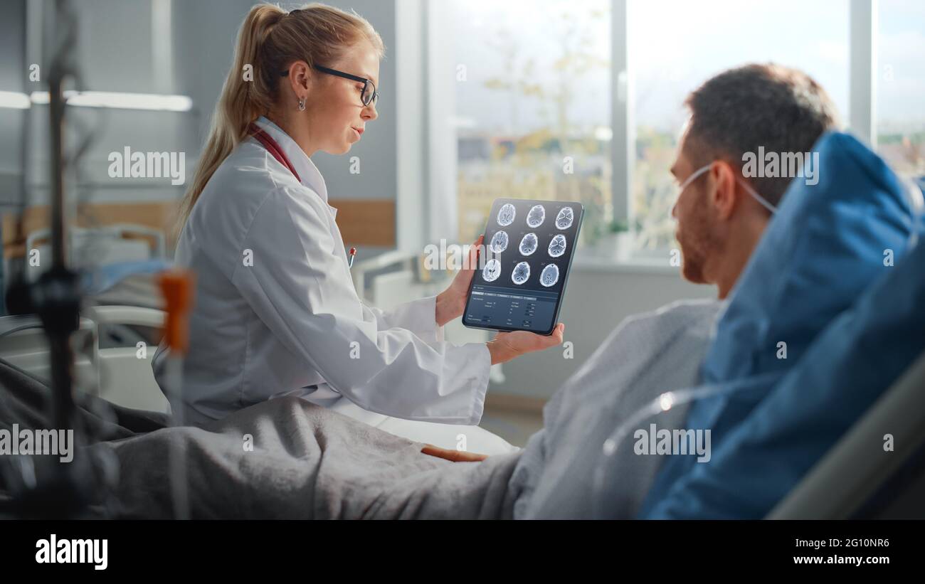 Krankenhausabteilung: Freundliche Ärztin spricht mit kranken männlichen Patienten, die im Bett ruhen, nutzt Tablet-Computer, zeigt und erklärt die Ergebnisse des CT-MRT-Scans des Gehirns Stockfoto