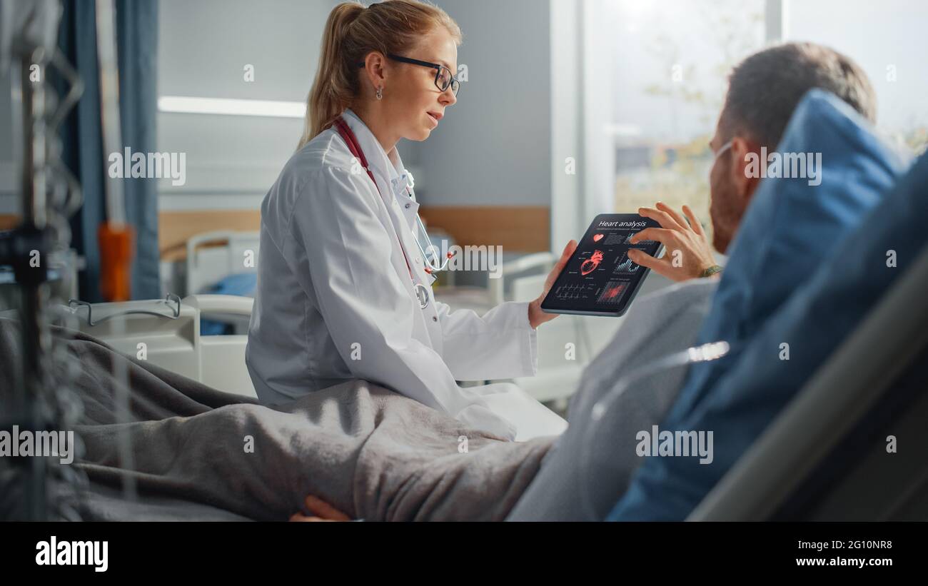 Krankenhausabteilung: Freundliche Ärztin spricht mit kranken männlichen Patienten, die im Bett ruhen, verwendet Tablet-Computer, zeigt und erklärt die Ergebnisse von Herz-Scans Stockfoto