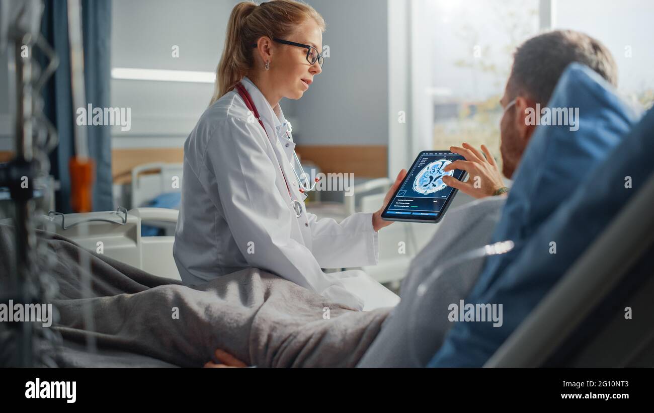 Krankenhausabteilung: Freundliche Ärztin spricht mit kranken männlichen Patienten, die sich im Bett ausruhen, verwendet einen Tablet-Computer, zeigt und erklärt die Ergebnisse von Gehirnscans Stockfoto