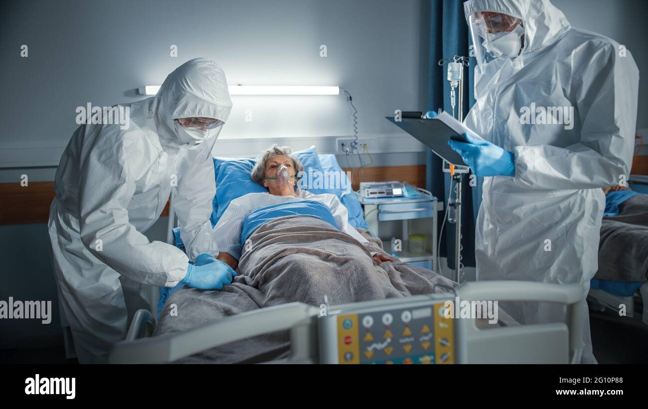 Krankenhaus Coronavirus Notaufnahme Station: Doktor trägt Overall schreibt unten die Vitalparameter einer älteren weiblichen Patientin mit Sauerstoffmaske, die im Bett ruht Stockfoto