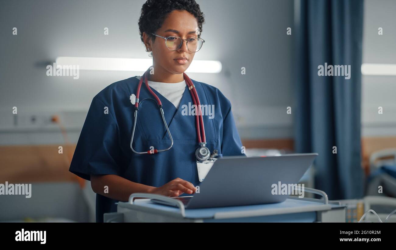 Krankenstation: Professionelle lächelnde schwarze weibliche Kopf Krankenschwester oder Doktor trägt Stethoskop verwendet medizinischen Computer. Im Hintergrund Patienten in Betten Stockfoto