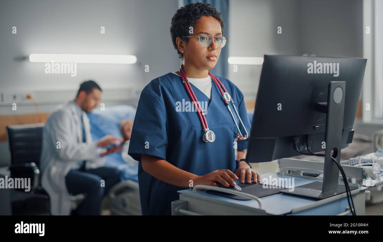 Krankenstation: Professionelle lächelnde schwarze weibliche Kopf Krankenschwester oder Doktor trägt Stethoskop verwendet medizinischen Computer. Im Hintergrund Patienten in Betten Stockfoto