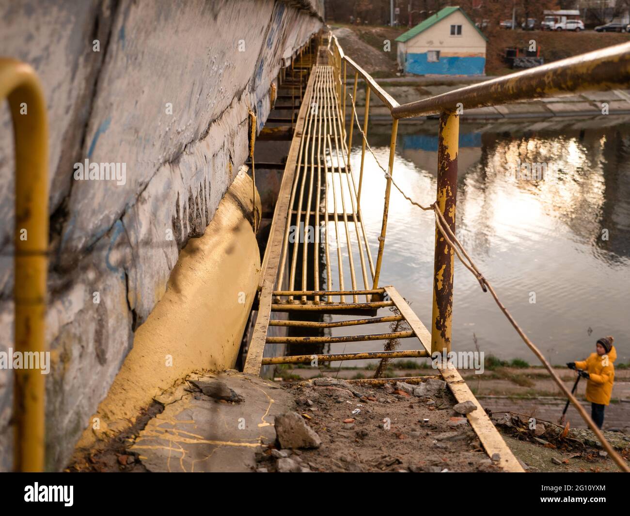 Schmale technische Brücke mit Metallstangen für die Instandhaltung einer großen Betonbrücke über den Wasserkanal. Boy mit dem Kick Scooter unter der Brücke. Stockfoto