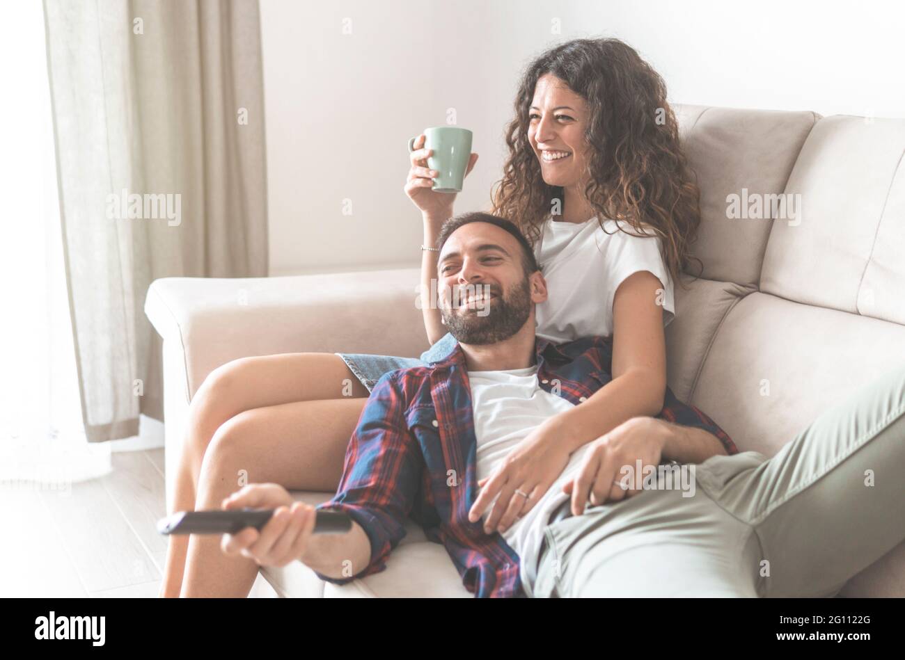 Junges Paar, das auf dem Sofa sitzt, Fernsehen schaut und eine Tasse Kaffee trinkt - Liebhaber entspannen sich und genießen Freizeit zu Hause Stockfoto