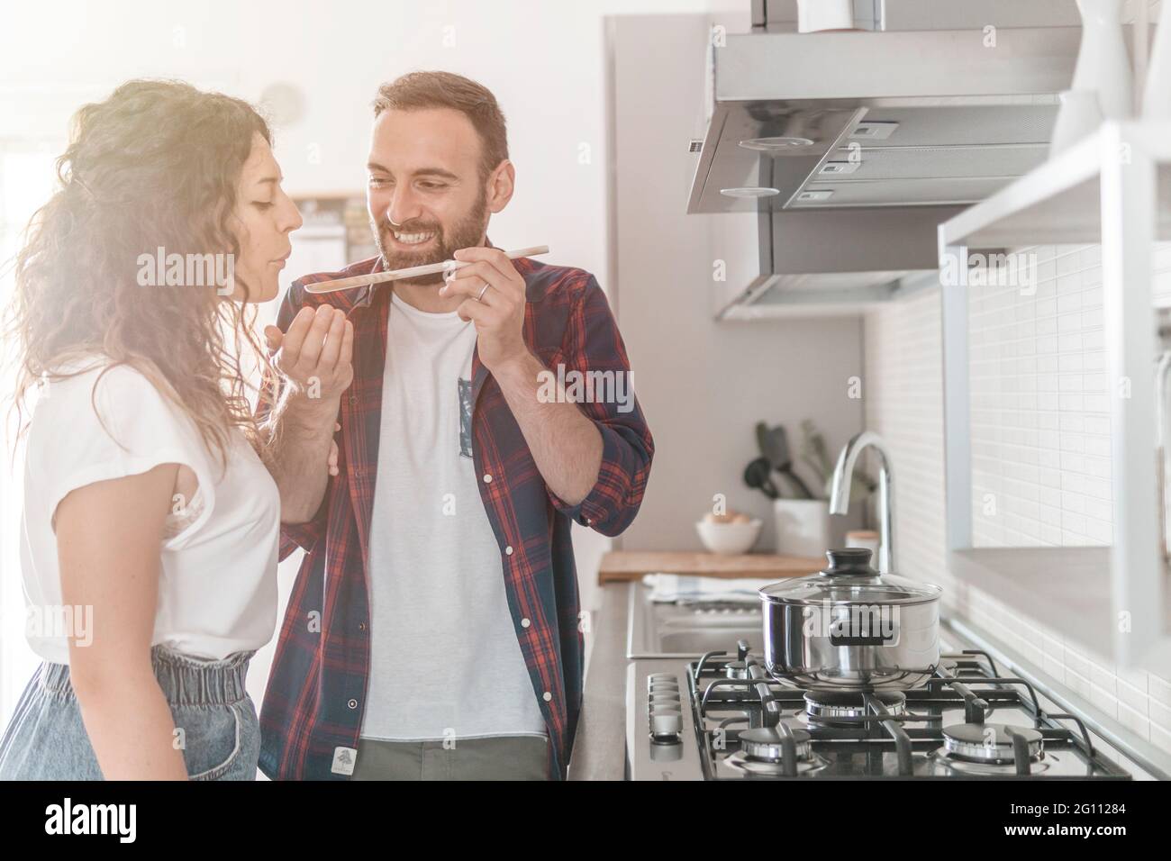 Das schöne junge Paar lächelt, während es das vorbereitete Essen kostet - das Paar hat Spaß in seiner neuen Küche, die Gerichte zubereitet Stockfoto
