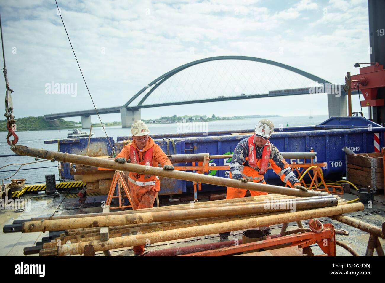 Fehmarn, Deutschland. Juni 2021. Premslaw Niecieki (l-r) und der Bohrtechniker Sergej Reinhardt demontieren während der Probebohrungen im Fehmarn Sound die Stangen einer Bohrung auf einem Bohrgerät. Mit den Bohrungen wird der Bau des Fehmarsundtunnels zwischen dem Schleswig-holsteinischen Festland und der Insel Fehmarn geplant. Im Hintergrund ist die Fehmarsundbrücke zu sehen. Quelle: Gregor Fischer/dpa/Alamy Live News Stockfoto