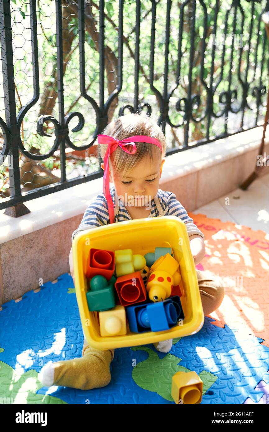 Das kleine Mädchen mit einer Schleife auf dem Kopf sitzt auf einem Teppich  auf dem Balkon und hält eine Kiste Spielzeug in den Händen Stockfotografie  - Alamy