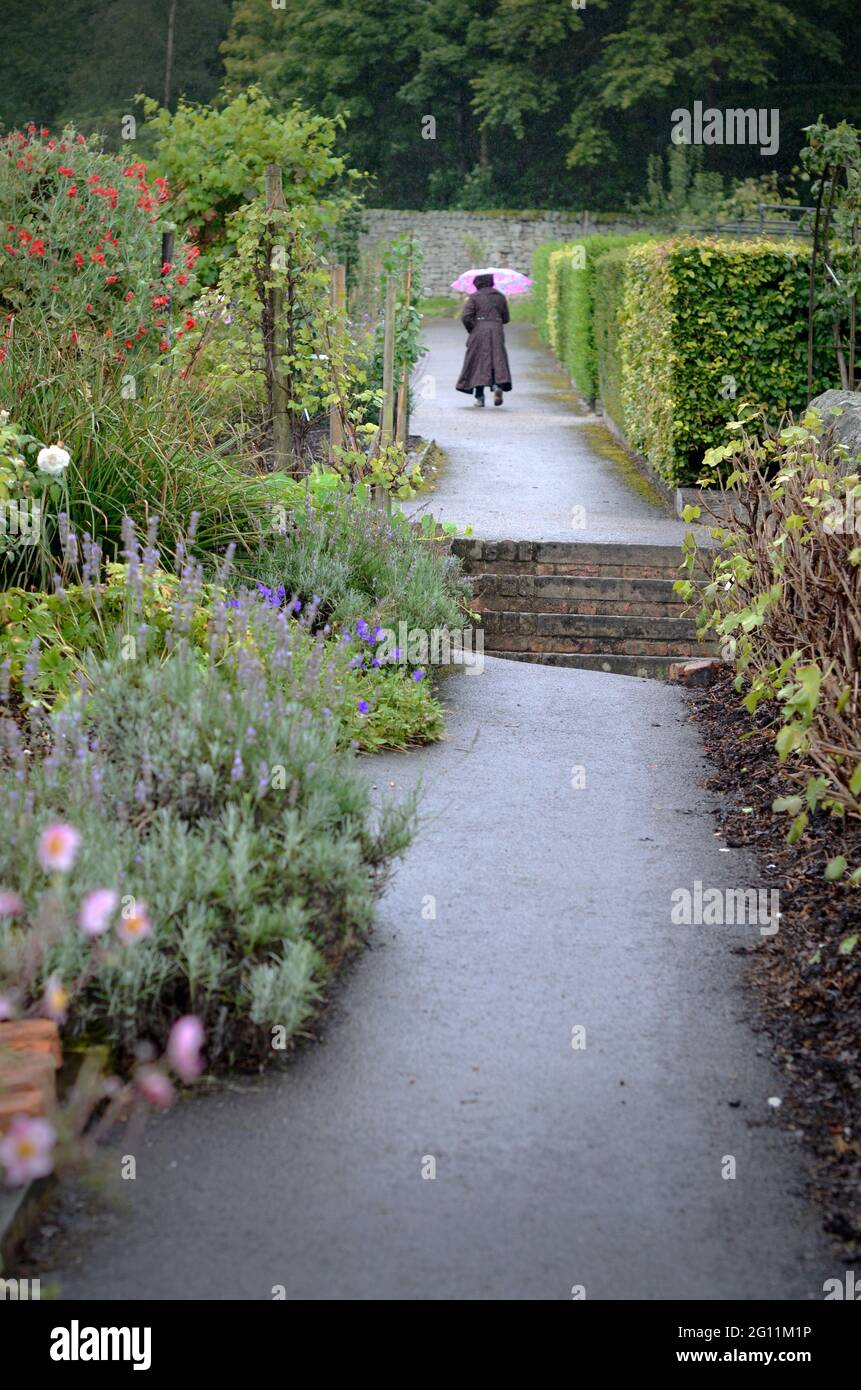 Einsame Frau, die im Regen mit einem Regenschirm durch den formellen Garten läuft Stockfoto