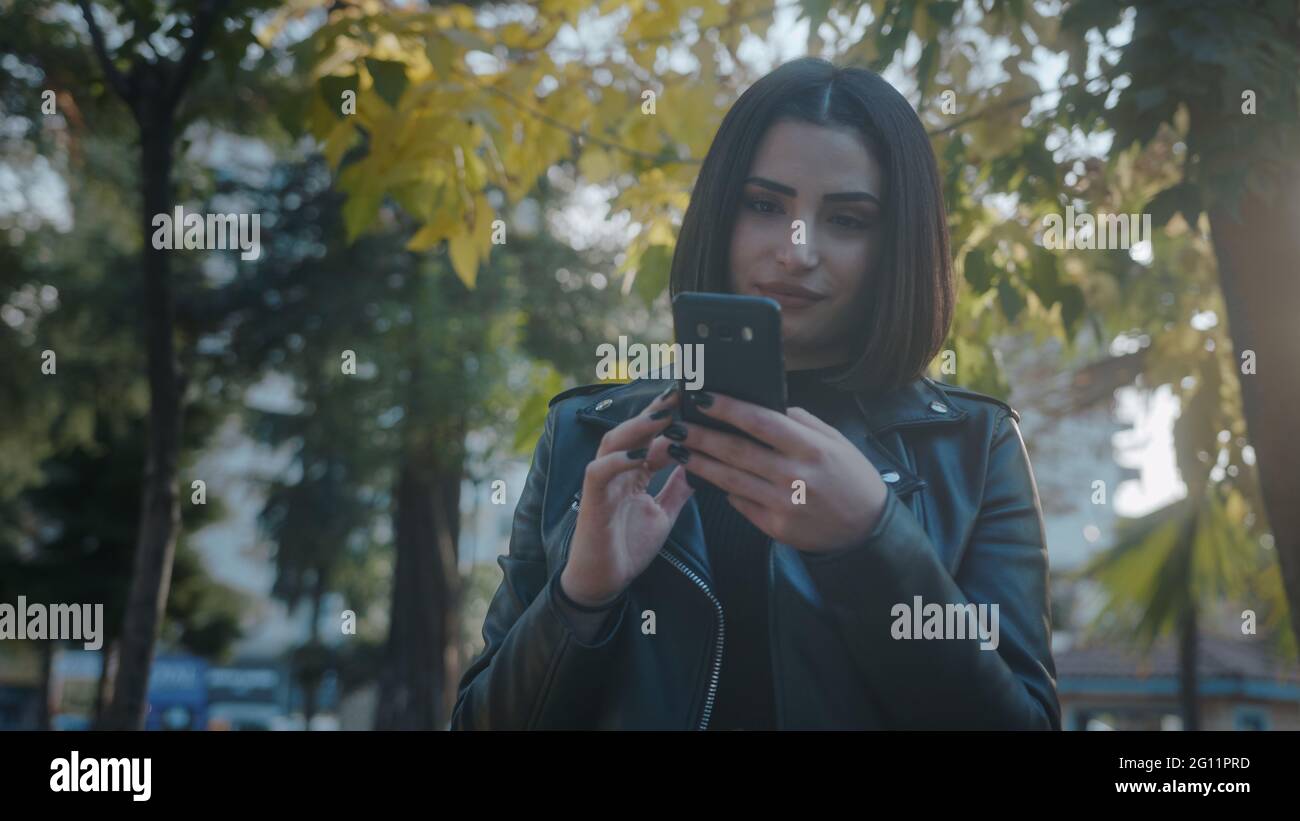 Schöne junge Frau mit Lederjacke lächelnd und tragen mit Handy im Park. Stockfoto