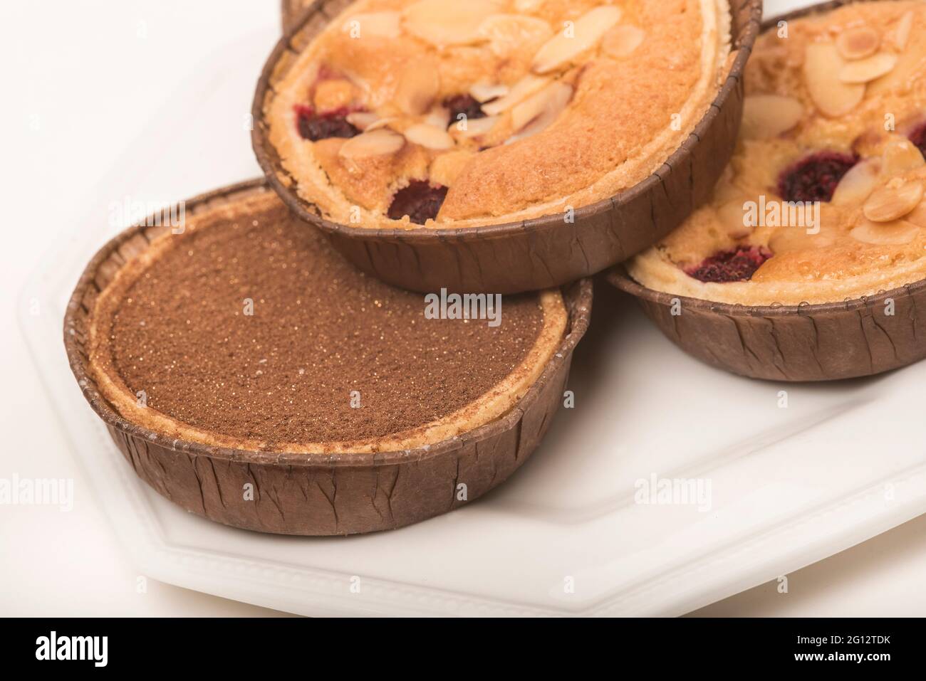 Himbeer-Bakewell und Schokolade und Toffee-Torten mit zwei dekorierten Tassen Tee/Kaffee für den Nachmittagstee. Weißer Hintergrund. Gebäck. Stockfoto