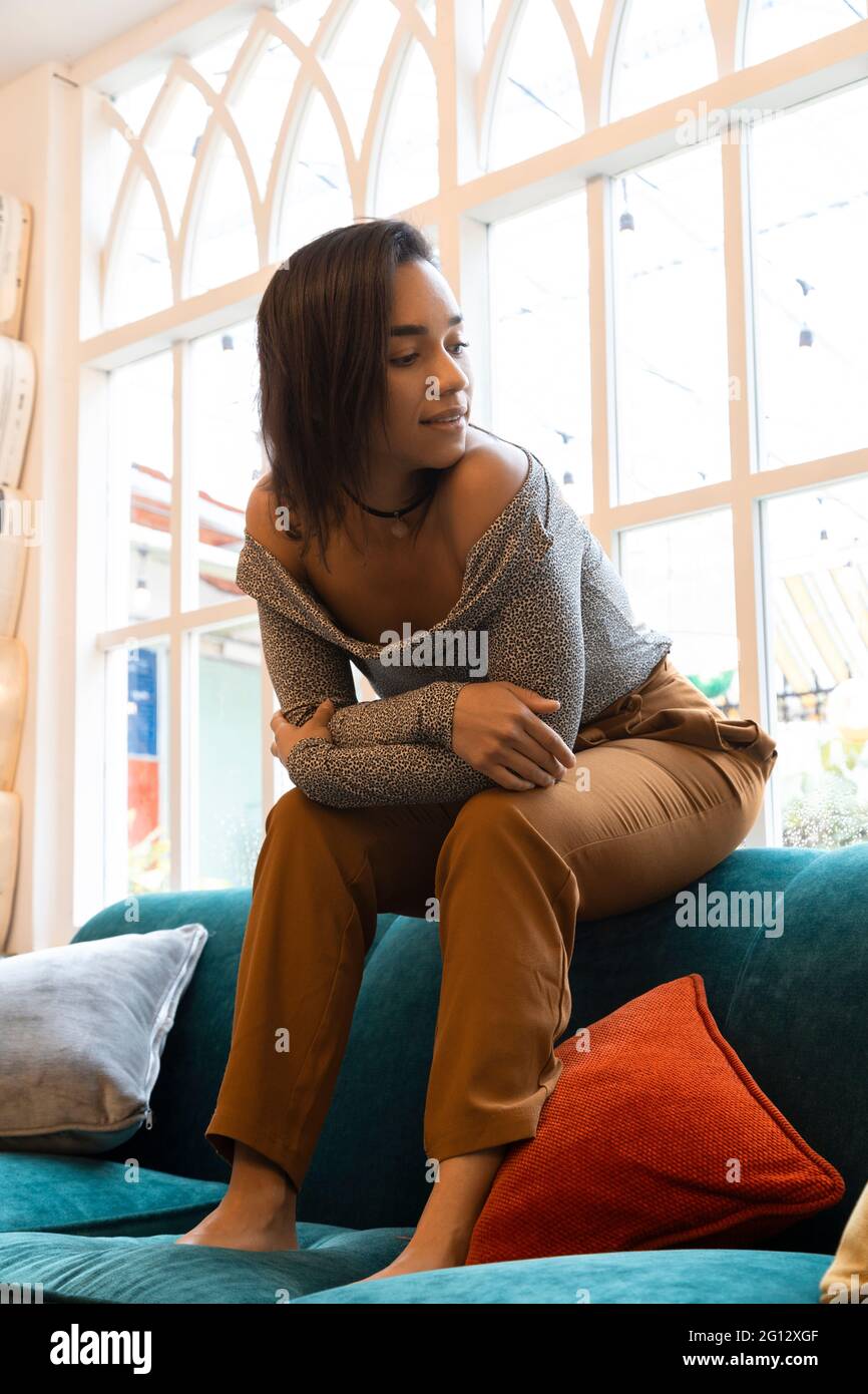 Lustige Brünette Frau, junge Latina mit kurzen Haaren am Rand eines bunten Sofas sitzend, bequeme Kleidung tragend, entspannter Lebensstil zu Hause Stockfoto